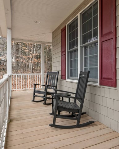 Covered porch with two black rocking chairs, wood decking, and view of bare trees through railing