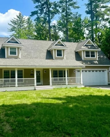 Two-story colonial house with three dormers, white railings, front porch, and attached garage surrounded by tall trees and green lawn.