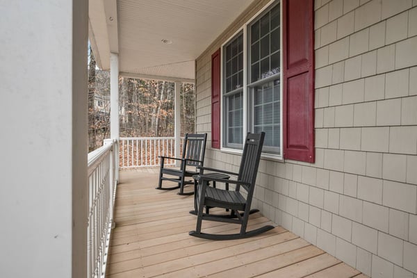 Covered porch with two black rocking chairs, wood decking, and view of bare trees through railing