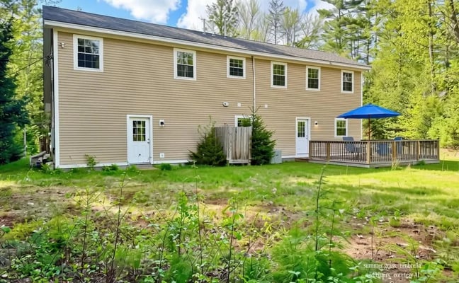 Two-story tan house with white trim and dark roof, wooden deck with blue umbrella, surrounded by green lawn and pine trees.