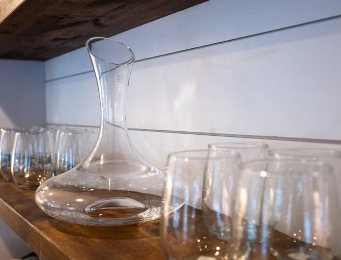 Glass decanter and drinking glasses arranged on wooden shelf against white wall